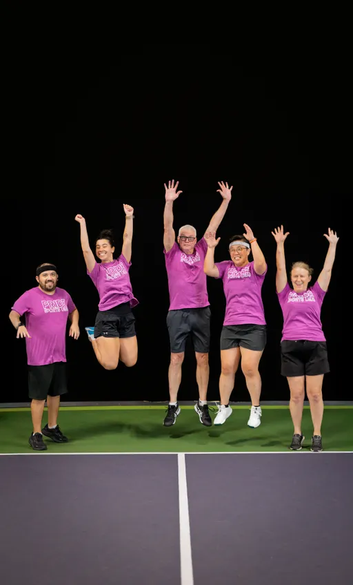 Five pickleball players wearing matching bright purple "Pride Pickleball" shirts and black athletic shorts are celebrating on a pickleball court, with four players mid-jump and all raising their arms high in the air. The group is positioned on the court's baseline against a black background, appearing joyful and energetic.