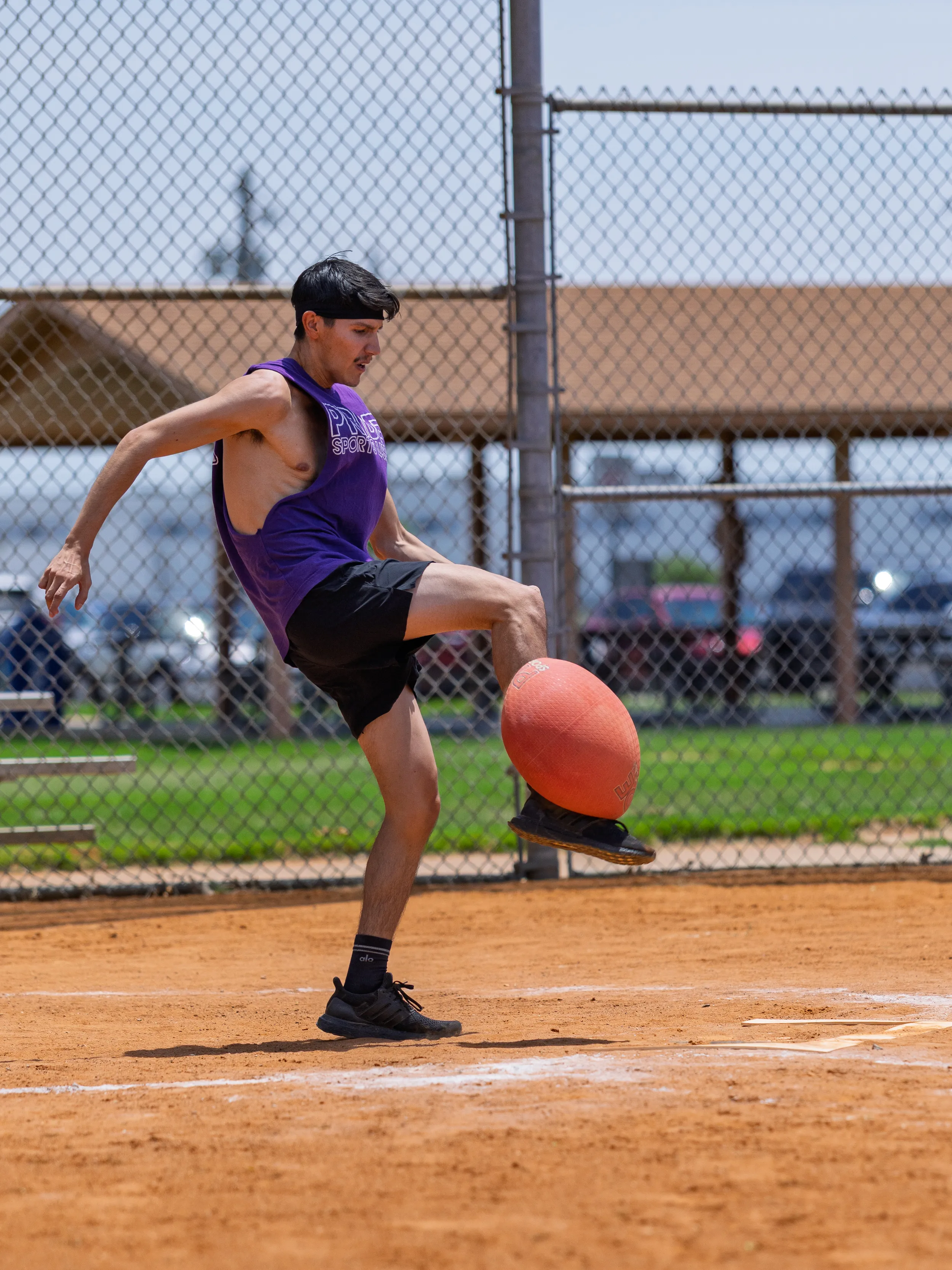 On a sunlit kickball field, a player in a purple “PRIDE” tank top, black shorts, and black sneakers lifts a leg to strike a bright red kickball near home plate, with a chain-link fence, shaded benches, and grassy outfield in the background.