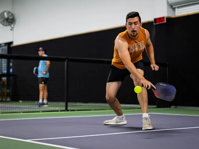 A player wearing a "PRIDE" tank top and shorts lunges forward on an indoor pickleball court, reaching out with a paddle to hit a pickleball, while another player stands in the background near the net.