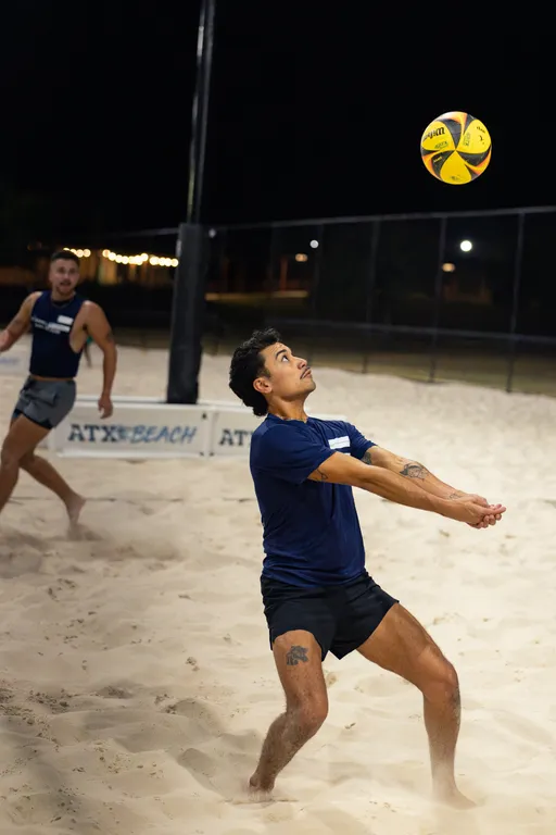 On a lit sand volleyball court at night, a player in a navy shirt and black shorts prepares to bump a yellow-and-blue volleyball, arms together and eyes up. A teammate in a matching top moves behind near the net and an “ATX BEACH” barrier, with fencing and park lights in the background.
