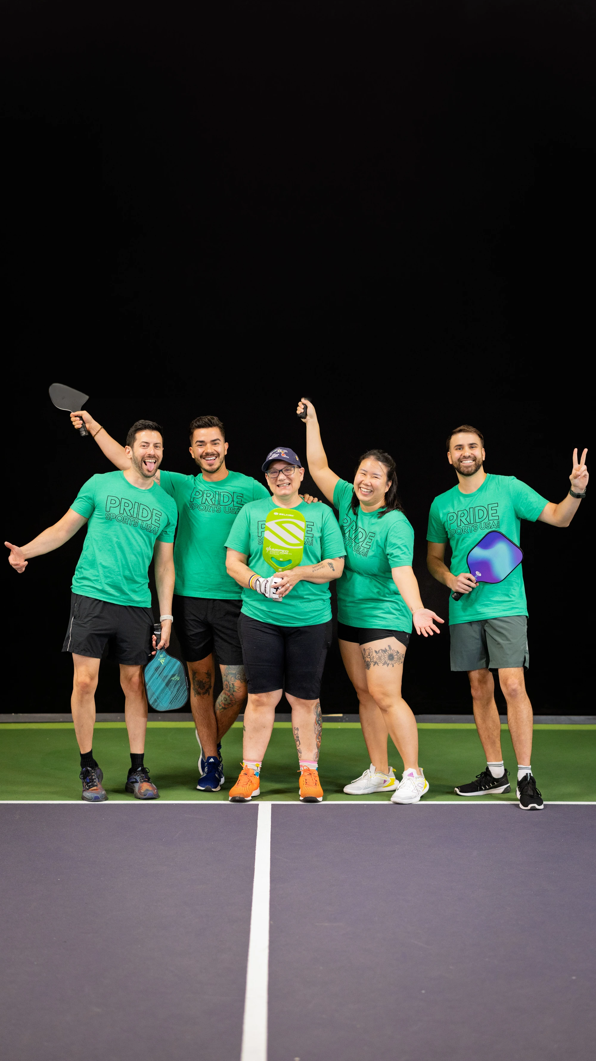 Five pickleball players stand together on an indoor court, all wearing matching green “Pride” shirts and holding pickleball paddles while smiling and posing enthusiastically for the camera.