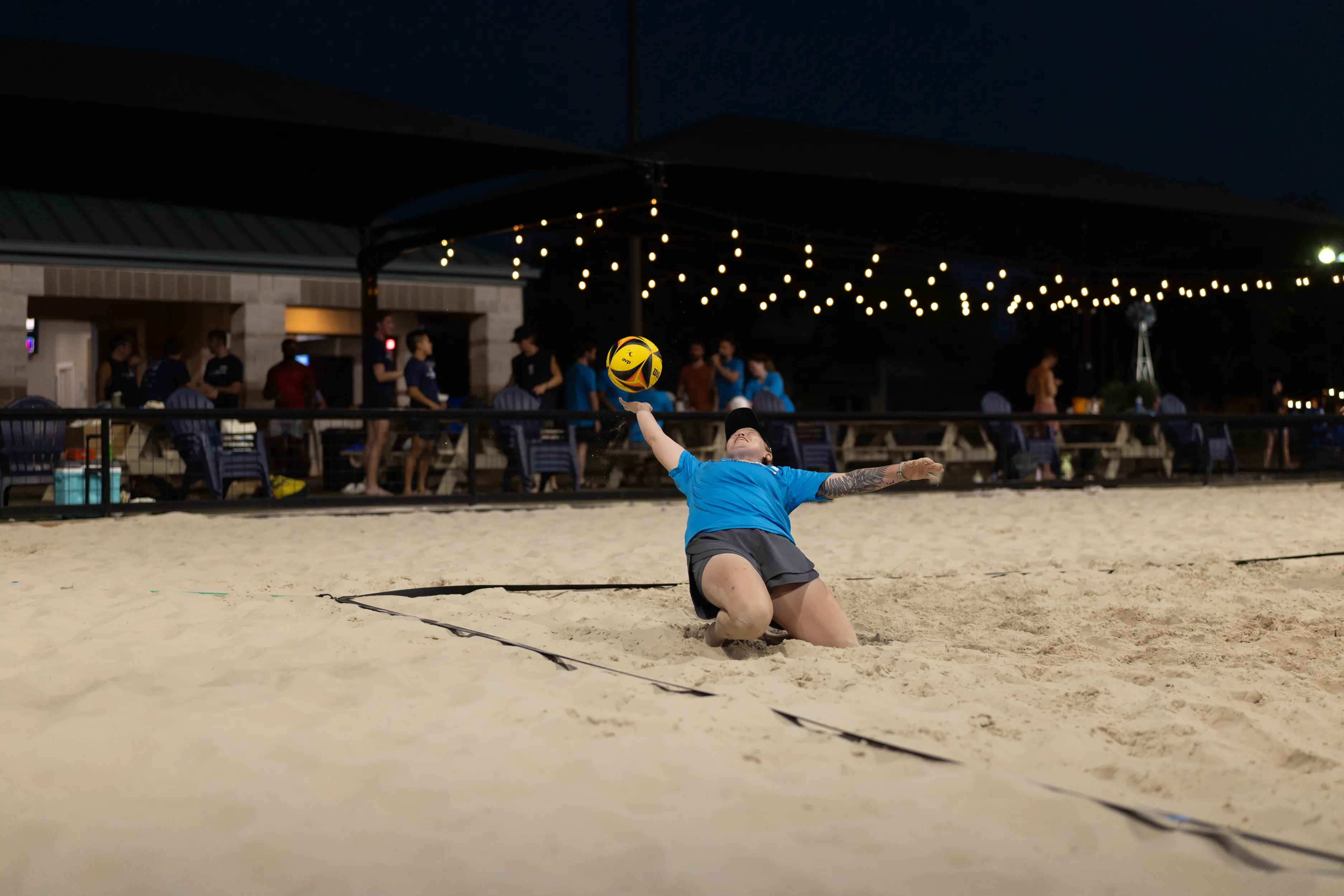 During a night match, a player in a blue shirt and gray shorts slides backward in the sand, reaching up with one hand to keep a yellow-and-black volleyball in play. String lights glow over the sand court as teammates and spectators watch from a pavilion and along the fence in the background.