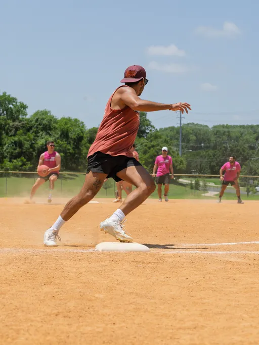 A kickball player wearing a sleeveless shirt, shorts, cap, and sunglasses is running past a base, while several competitors in matching pink shirts prepare to field the kickball on a sunny outdoor field.