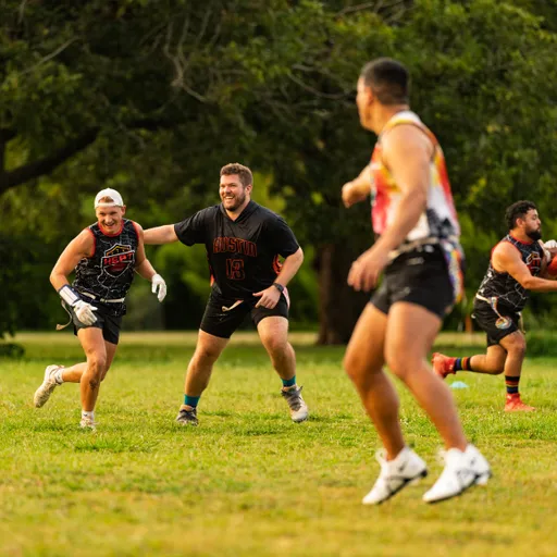 On a tree-lined park field, ATX Heat (Orange) players run flag football drills for Gay Bowl 2025, wearing black-and-orange jerseys (including an “Austin” top), multicolor practice tops, flag belts, gloves, and cleats; a smiling player cuts across the grass as teammates pursue and a runner blurs in the foreground.