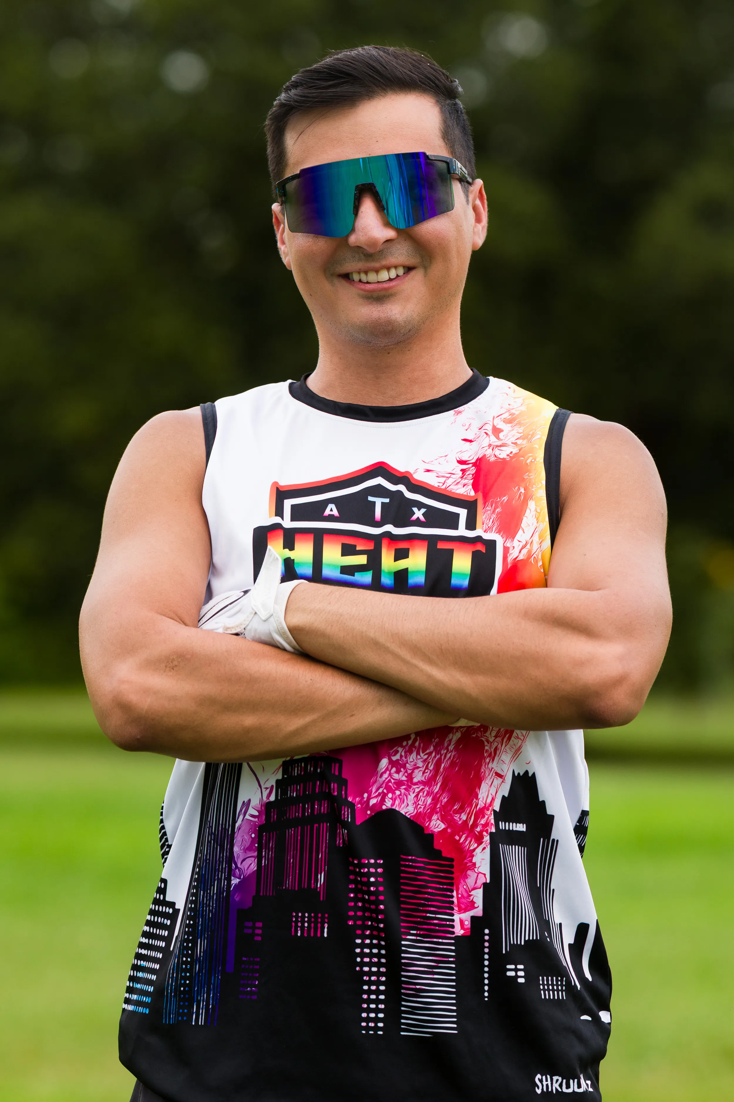 An ATX Heat player stands on a grassy practice field with arms crossed, wearing a sleeveless ATX Heat jersey with a colorful skyline and orange accent, reflective wraparound visor sunglasses, and a white football glove. Trees blur in the background, suggesting a relaxed portrait during Gay Bowl practice.