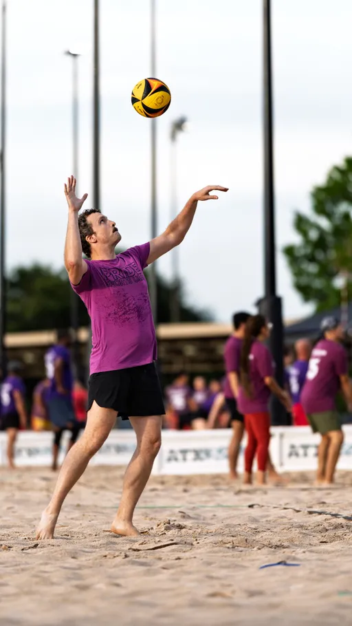A volleyball player wearing a purple "Pride" shirt and black shorts is in the process of serving a yellow and black volleyball on a sandy court, with other players and a net visible in the background.