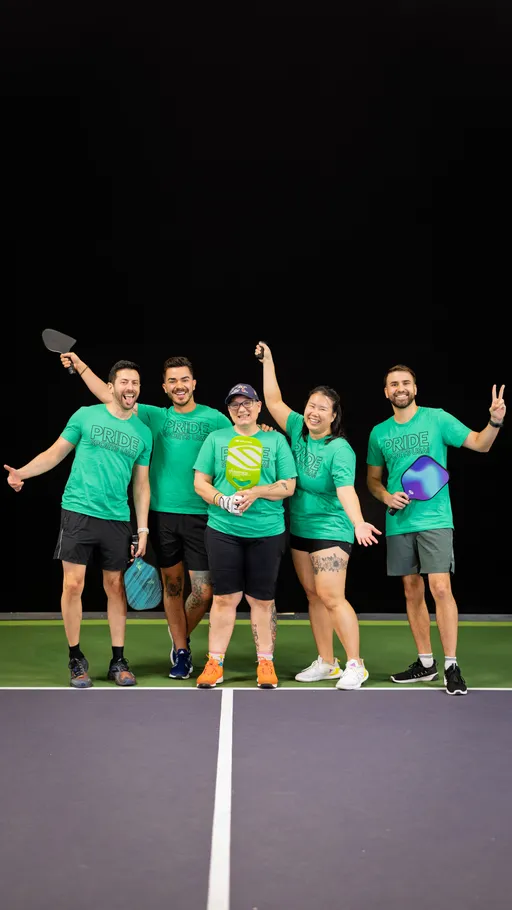 Five pickleball players stand together on an indoor court, all wearing matching green “Pride” shirts and holding pickleball paddles while smiling and posing enthusiastically for the camera.
