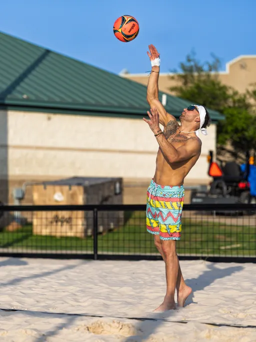 A volleyball player wearing colorful patterned shorts, sunglasses, and a white headband is in the motion of serving the ball on a sand court during a sunny day. The background features a green-roofed building, a fence, and outdoor equipment.