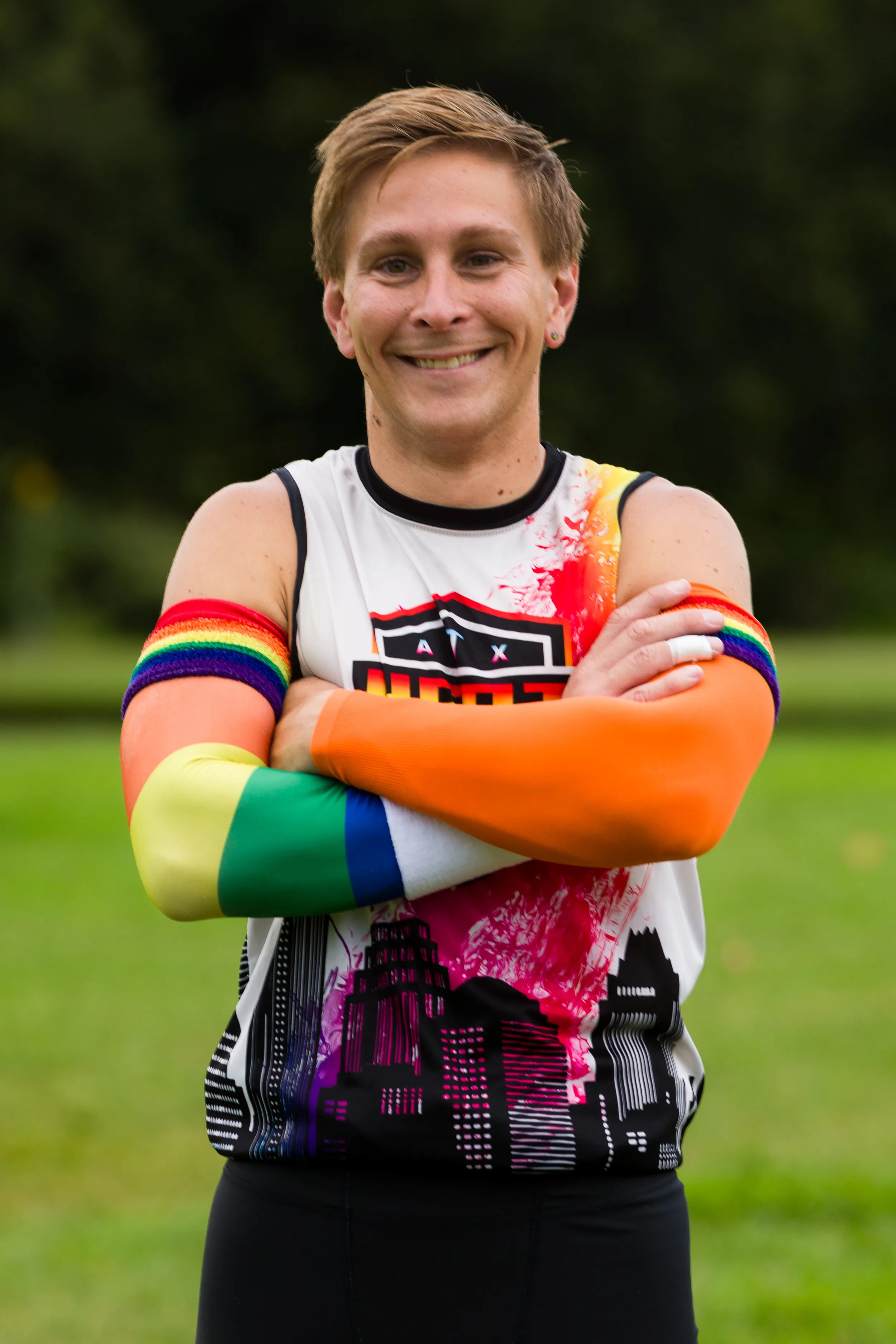 An ATX Heat (Orange) player stands on a grassy practice field with arms crossed, wearing a white sleeveless team jersey featuring the ATX Heat logo and a city skyline graphic, plus rainbow-striped and orange compression arm sleeves, with trees in the background during Gay Bowl 2025 practice.
