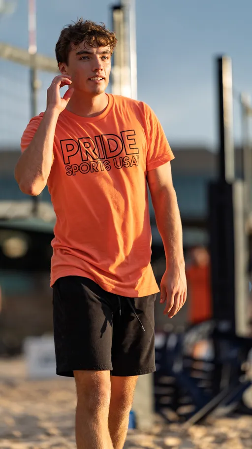 A volleyball player wearing a bright orange "Pride Sports USA" t-shirt and black shorts is walking on a sand court, with a volleyball net and equipment in the background.