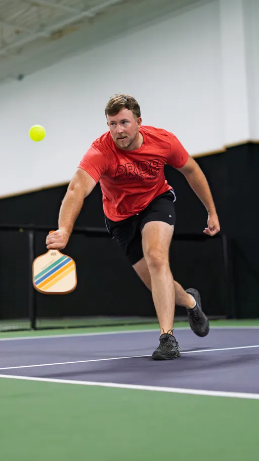 A pickleball player wearing a red "Pride" shirt and black shorts is lunging forward on an indoor court, reaching out with a paddle to return a ball in mid-air.