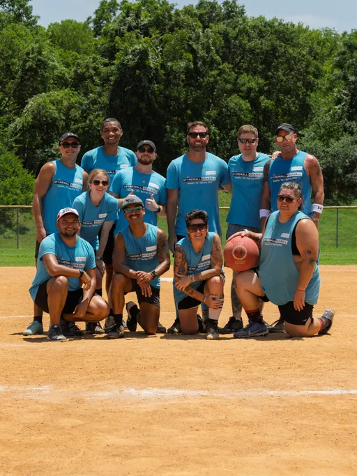 A group of kickball players pose together on a sunny kickball field, all wearing matching blue team shirts and athletic gear, with one player holding a red kickball. Trees and a fence are visible in the background.