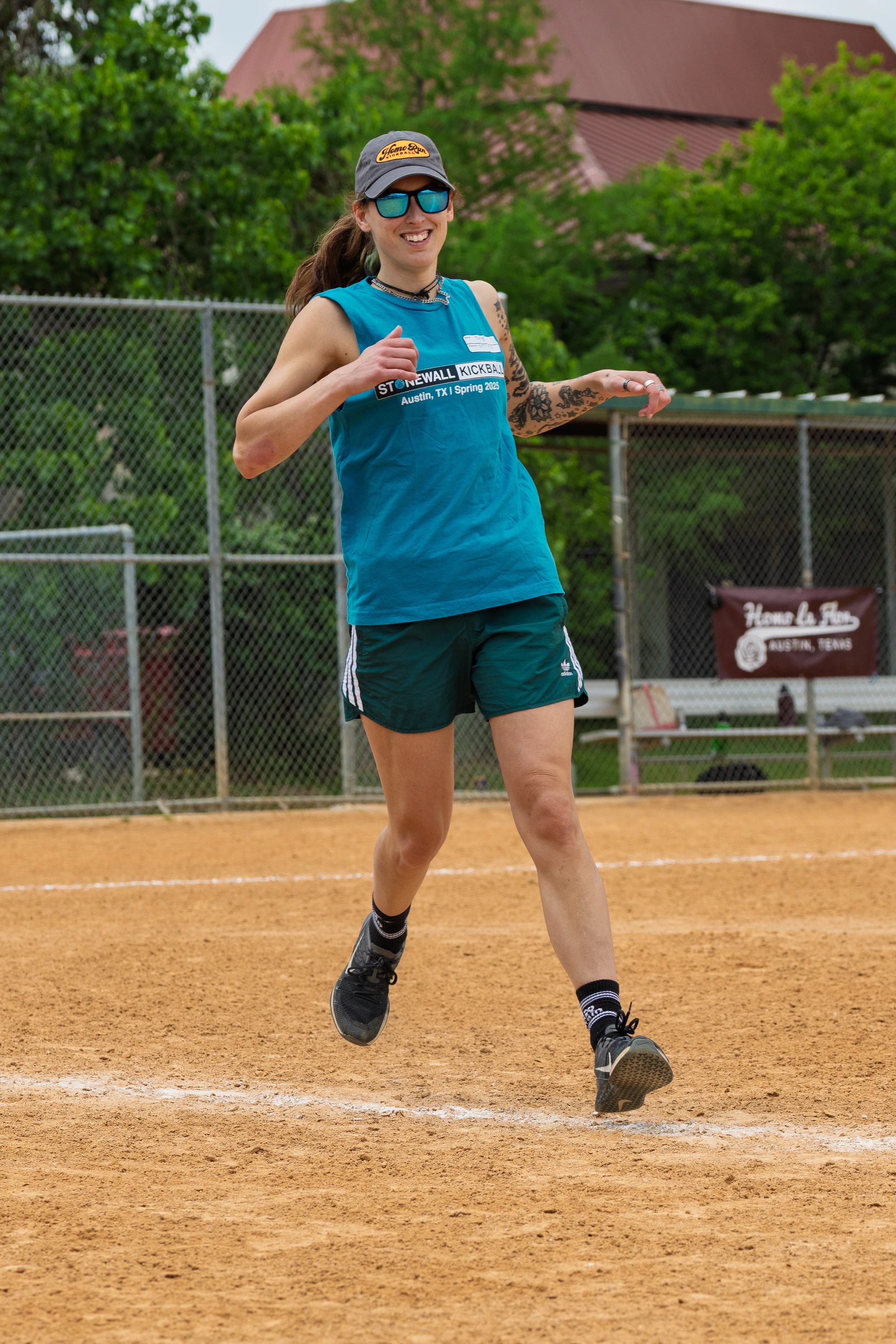 A player wearing a teal Stonewall Kickball tank top, dark athletic shorts, sunglasses, and a cap is running on a kickball field, appearing to celebrate or cheer with thumbs up. A chain-link fence and greenery are visible in the background.
