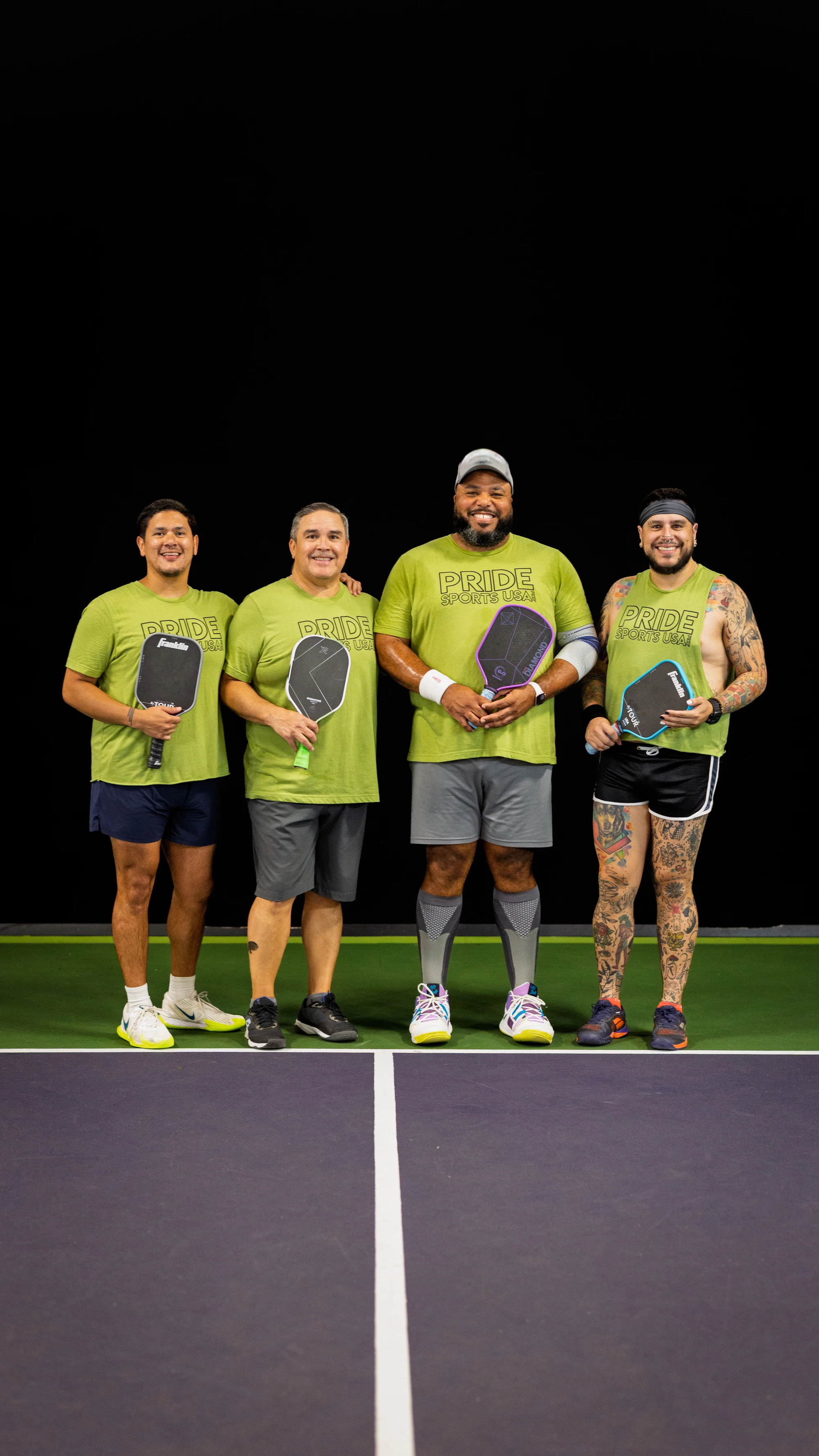 Four pickleball players stand side by side on an indoor pickleball court, each holding a paddle and wearing matching green "Pride Pickleball" shirts, with two players wearing wristbands and one wearing a cap. The group is smiling and posing for a team photo, and one player has visible tattoo sleeves and athletic shorts.