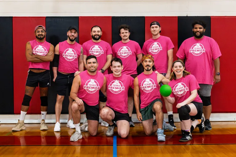 A group of dodgeball players poses for a team photo in an indoor gym, wearing matching pink “Texas Premier” shirts with athletic shorts and sneakers; some have knee pads, hats, or headbands. Several kneel in front—one holding a bright green dodgeball—against a red-and-black padded wall.