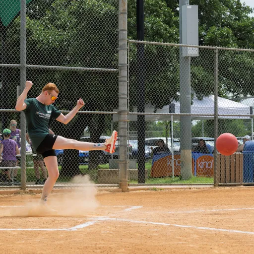 A player wearing a dark athletic shirt, shorts, and sunglasses is kicking a red kickball on a dirt field, with a cloud of dust rising from their foot and a chain-link fence and spectators in the background.