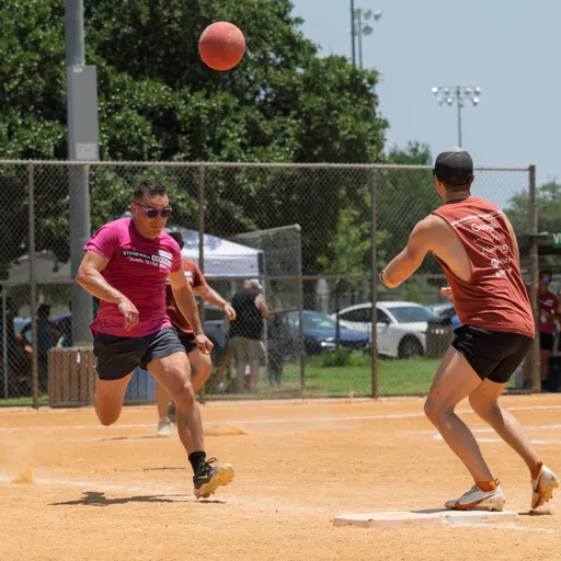 A player in a maroon shirt and athletic shorts is running toward a base as another player in a red tank top stands ready to catch the kickball, which is in mid-air above the base on a dusty kickball field with a fence and trees in the background.