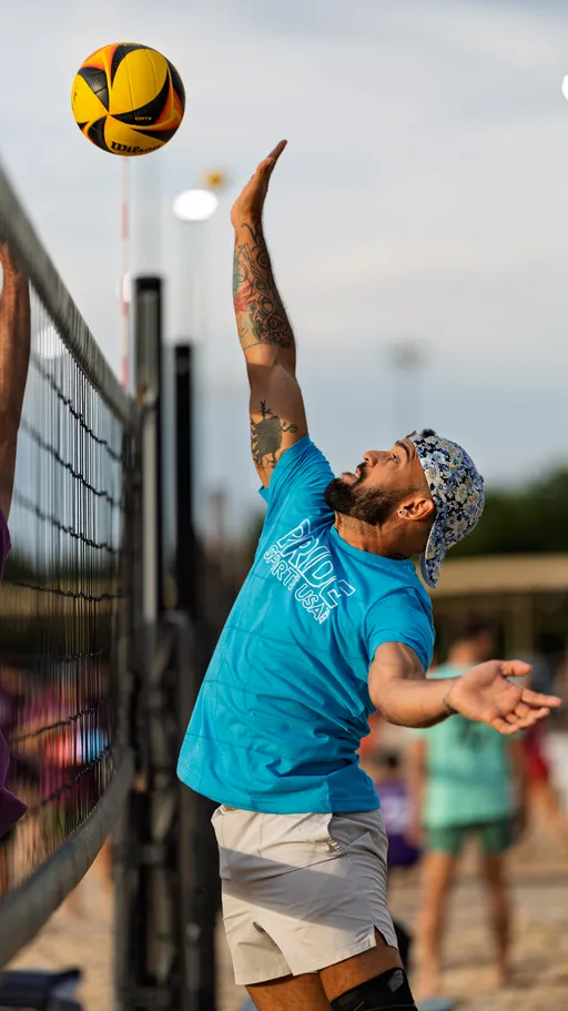 A volleyball player wearing a blue "Pride Sports USA" t-shirt, light shorts, and a patterned cap jumps at the net with an outstretched arm, preparing to hit a yellow and black volleyball during a sand volleyball game. Other players and a sandy court are visible in the background.