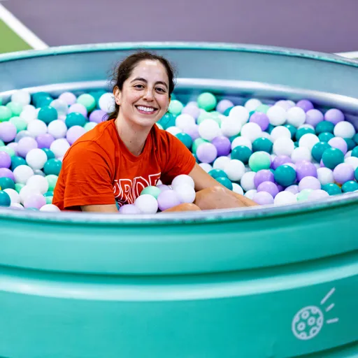 A player wearing a bright orange t-shirt sits and smiles in a turquoise ball pit filled with pastel-colored plastic balls, positioned next to a pickleball court.