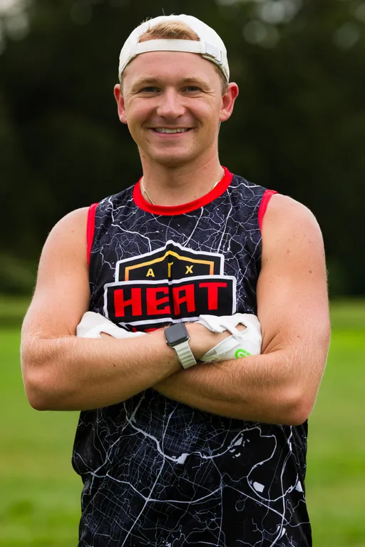 An ATX Heat player stands on a grassy practice field with arms crossed, smiling during Gay Bowl 2025 preparations. The athlete wears a black ATX HEAT sleeveless jersey with red trim, white football gloves, a backward white cap, and a smartwatch, with trees softly blurred in the background.