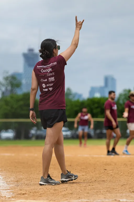 A player wearing a maroon team shirt, black shorts, and athletic shoes stands on a kickball field with one arm raised, signaling, while other team members are visible in the background preparing for play. The scene takes place outdoors with a city skyline blurred in the distance.
