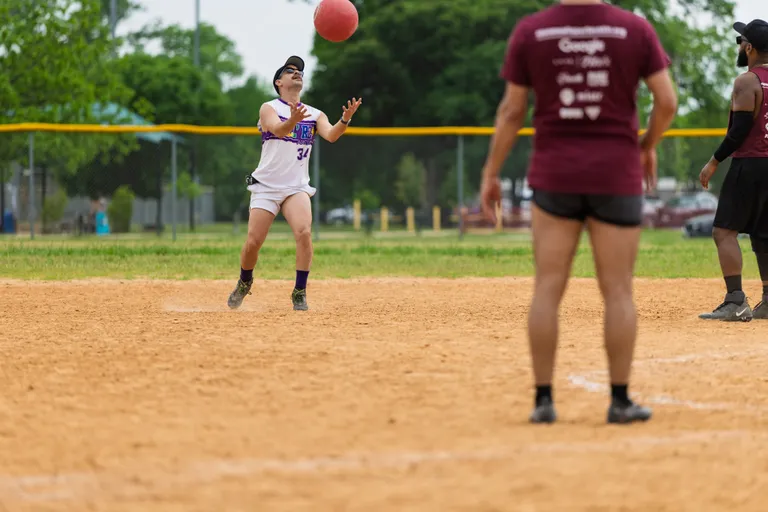 A player wearing a white uniform, black cap, and purple socks is preparing to catch a red kickball on a sandy kickball field, while competitors in maroon shirts watch from nearby.