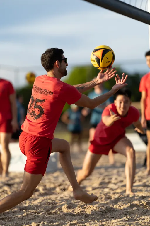 Two volleyball players wearing matching red shirts and shorts are competing on a sandy outdoor court, with one athlete reaching out and preparing to set or catch a yellow and black volleyball mid-air, while another player crouches and extends an arm in the background.