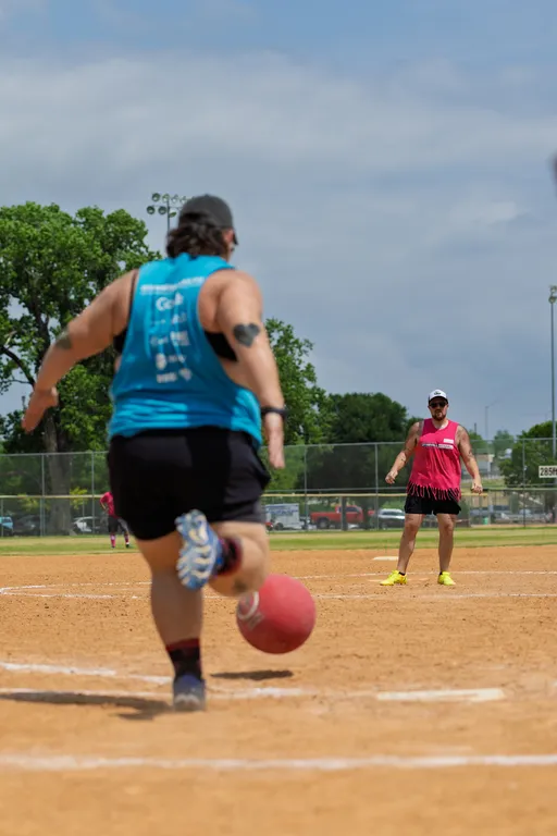 A player wearing a blue tank top and black shorts is running up to kick a red kickball on a dirt field, while another player in a pink fringed tank top and neon shoes stands ready in the background; the scene takes place outdoors with a chain-link fence and trees visible behind the field.
