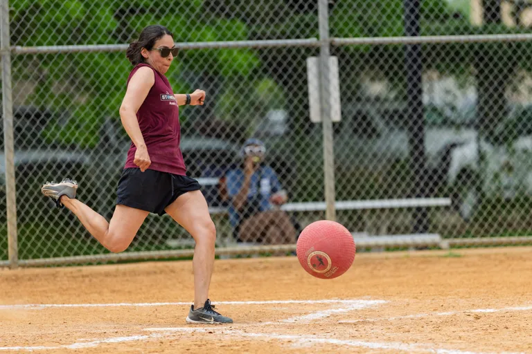 A player wearing a maroon sleeveless shirt, black shorts, and sunglasses is kicking a red kickball on a dirt field, with a chain-link fence and a seated person visible in the background.