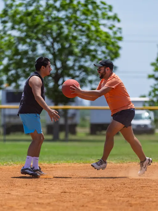Two players are on a kickball field; one is leaping forward holding a bright red kickball, while the other stands ready to receive or block, both dressed in athletic clothing and sneakers. Trees and a yellow field fence are visible in the background on a sunny day.