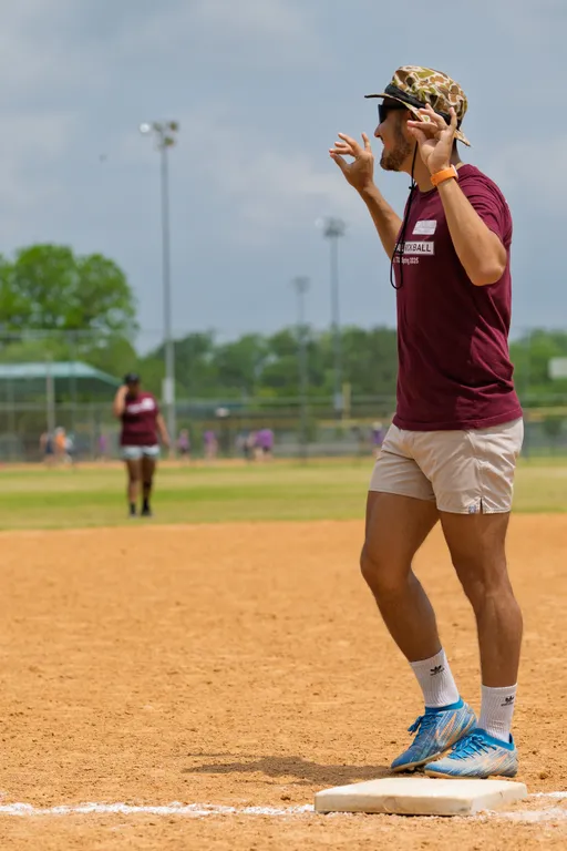 A kickball player wearing a maroon shirt, light shorts, blue athletic shoes, and a patterned hat stands on a base with their arms raised, appearing to react to the action on the field during a game on a sunny kickball field.