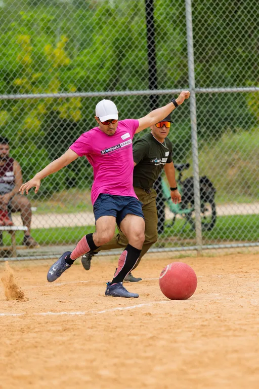 A player in a pink shirt, white cap, and sunglasses is kicking a red kickball on a sandy field while another competitor in a green shirt and sunglasses runs behind them, with a chain-link fence and greenery in the background.