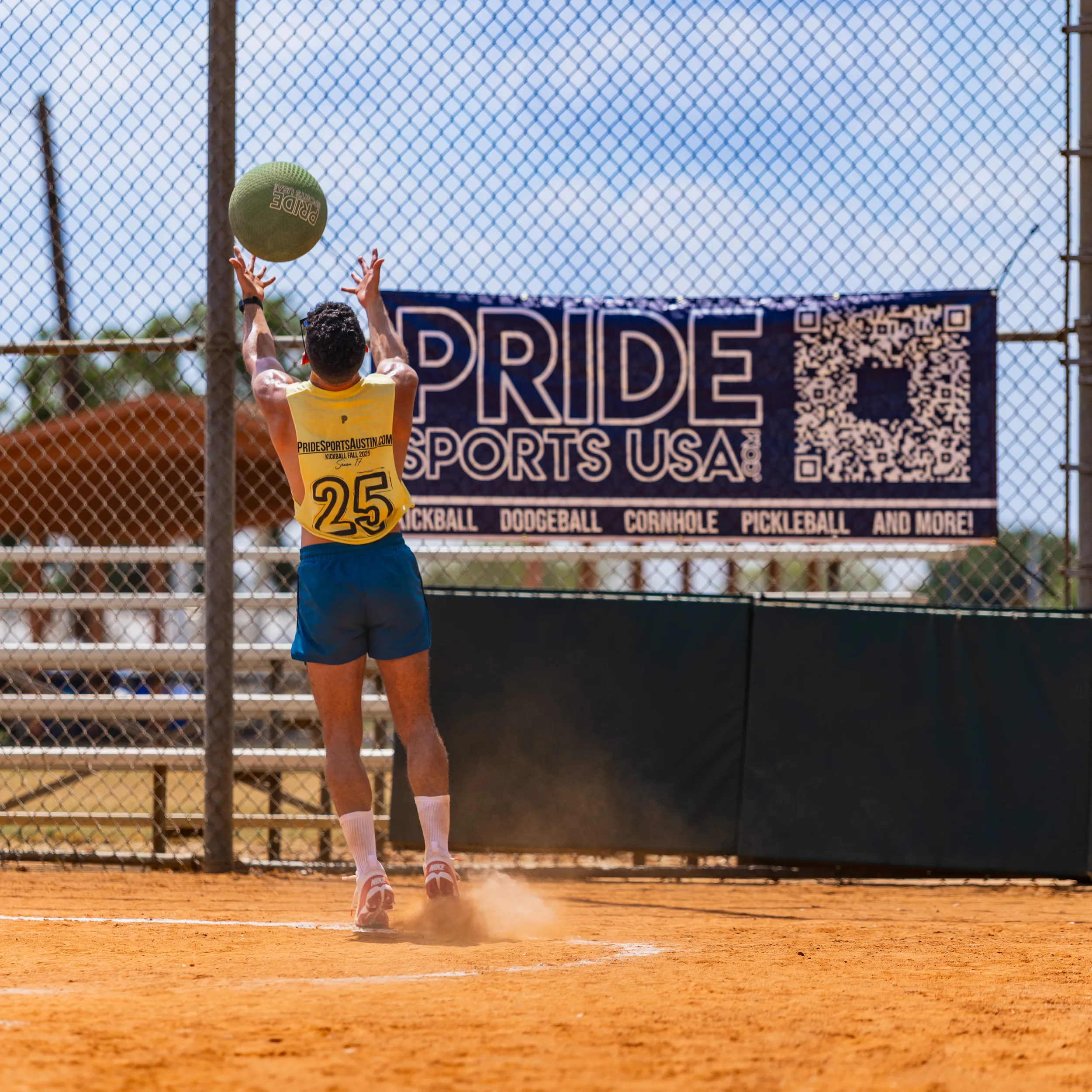 On a dusty kickball field, a Pitch Please player in a yellow jersey with number 25, blue shorts, and knee-high socks jumps at the backstop with arms raised to catch a green kickball, kicking up dust. A chain-link fence and a large PRIDE SPORTS USA banner fill the background in bright daylight.