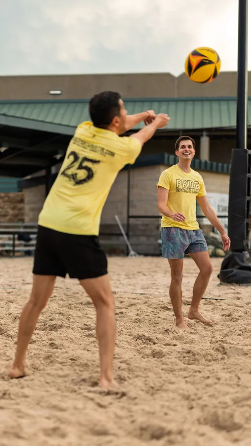 Two players wearing yellow Pride Sand Volleyball shirts and shorts are on a sandy volleyball court; one player is actively bumping the volleyball while the other looks on, ready to play.