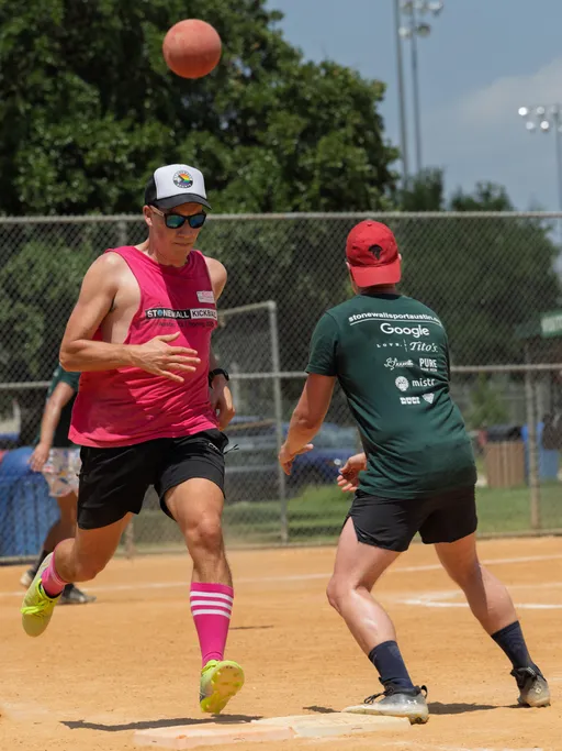 Two players are in action on a kickball field; one in a bright pink jersey and matching socks runs towards a base while another in a dark green shirt prepares to field the kickball that is mid-air above them. Both players wear athletic shorts and hats, and a chain-link fence and green trees are visible in the background.