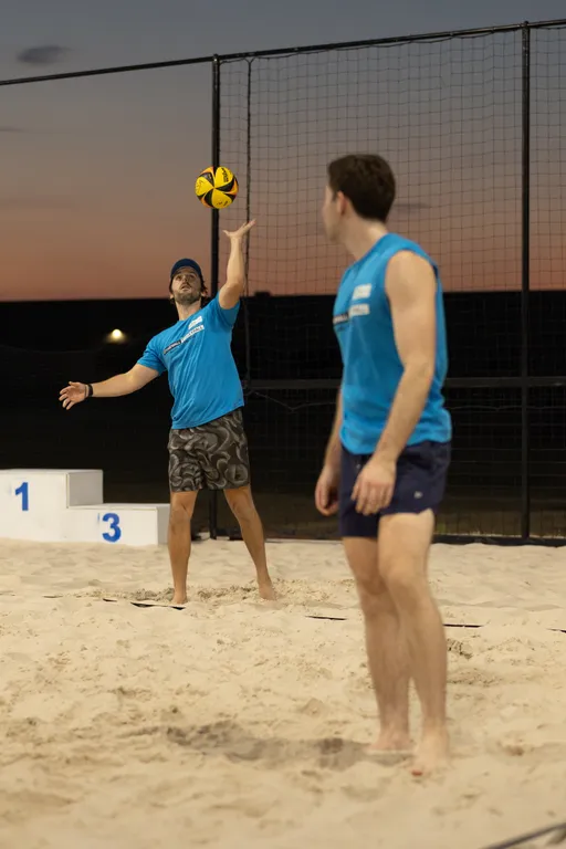 At dusk on a sand volleyball court, a player in a blue team shirt tosses a yellow-and-black ball to serve while a teammate in a matching top watches, ready to receive. The scene shows overhead lights, a tall safety net, a podium marked 1 and 3 in the background, and both athletes barefoot on the sand wearing shorts.