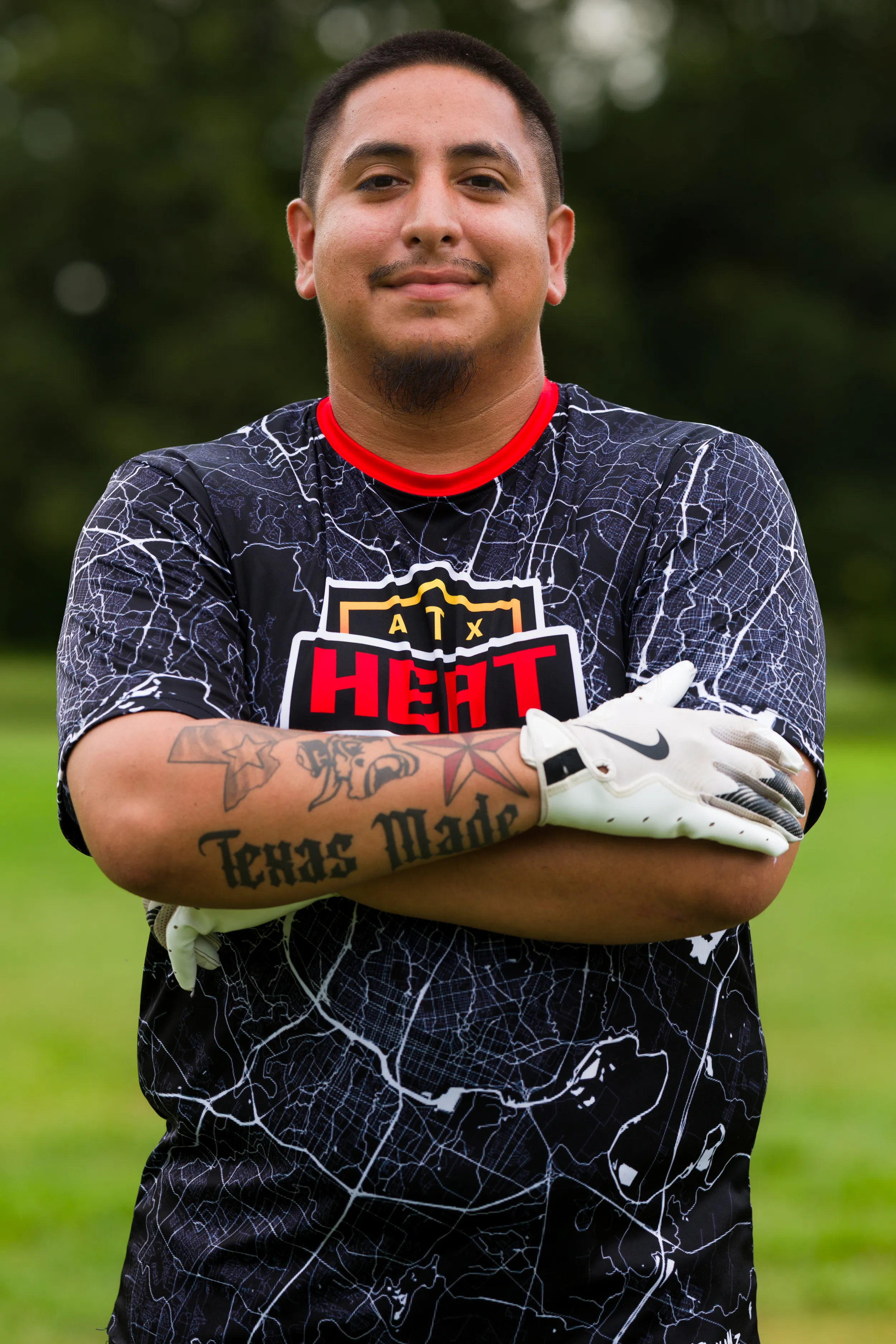 An ATX Heat player poses with arms crossed on a grassy field during Gay Bowl 2025 practice, wearing a black jersey with a white map pattern, red collar, and an “ATX HEAT” logo. The athlete has white football gloves and stands in an outdoor park setting with blurred trees in the background.