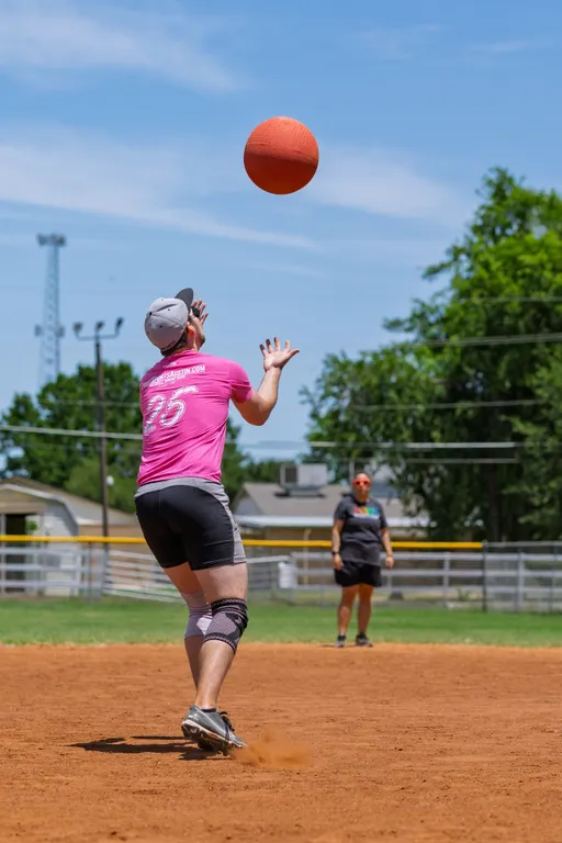 A kickball player wearing a pink jersey, black shorts, and a cap is preparing to catch a kickball in mid-air on a sunny outdoor field, while another player stands in the background near the outfield fence.
