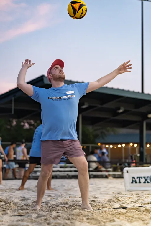 A volleyball player wearing a red cap, blue Stonewall Volleyball t-shirt, and shorts is preparing to serve the ball on a sandy outdoor court, with a pavilion and other participants in the background.
