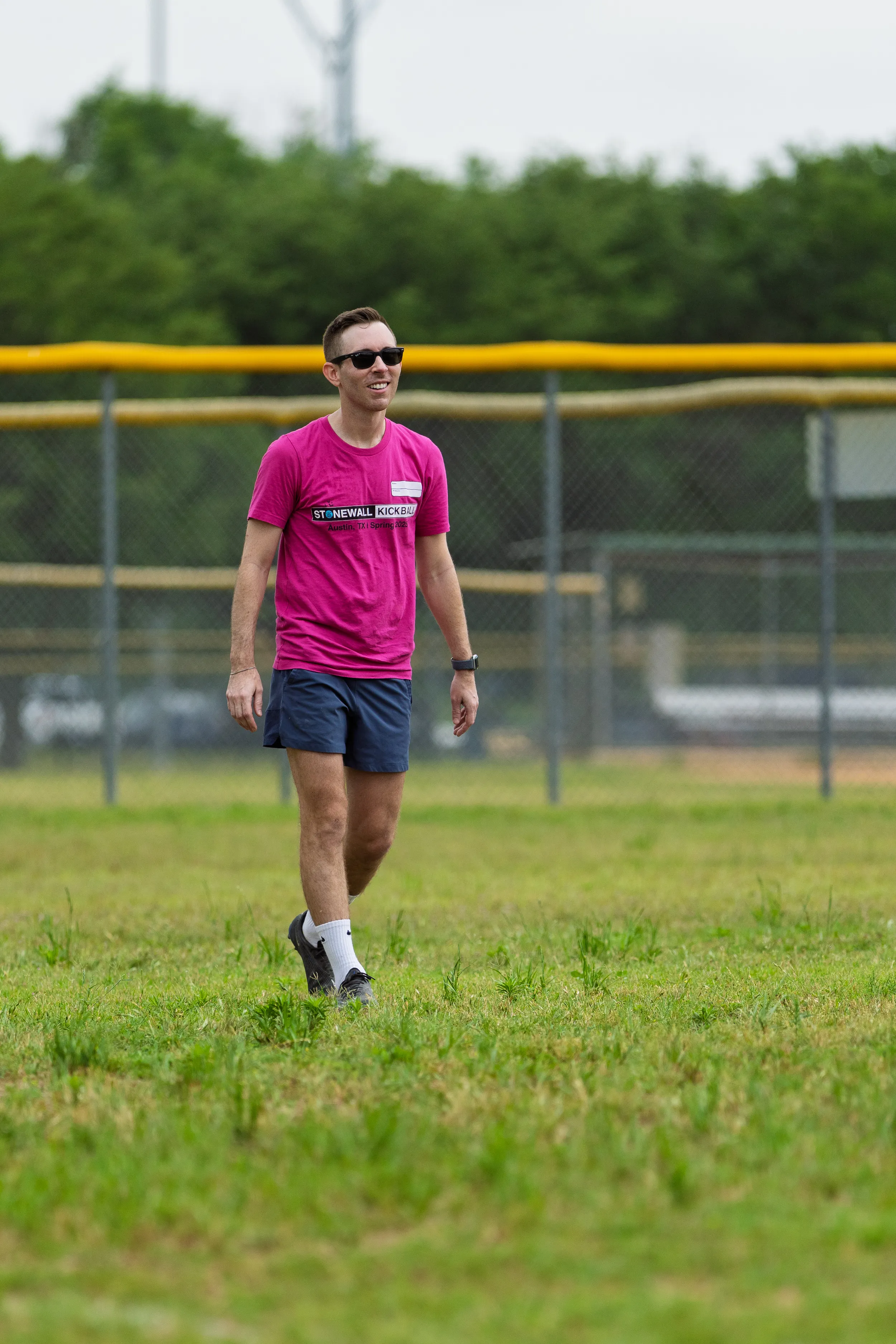 A player in a bright pink “STONEWALL KICKBALL” shirt, dark shorts, and sunglasses walks across a grassy kickball field, wearing white socks and athletic shoes, with chain-link fencing behind.