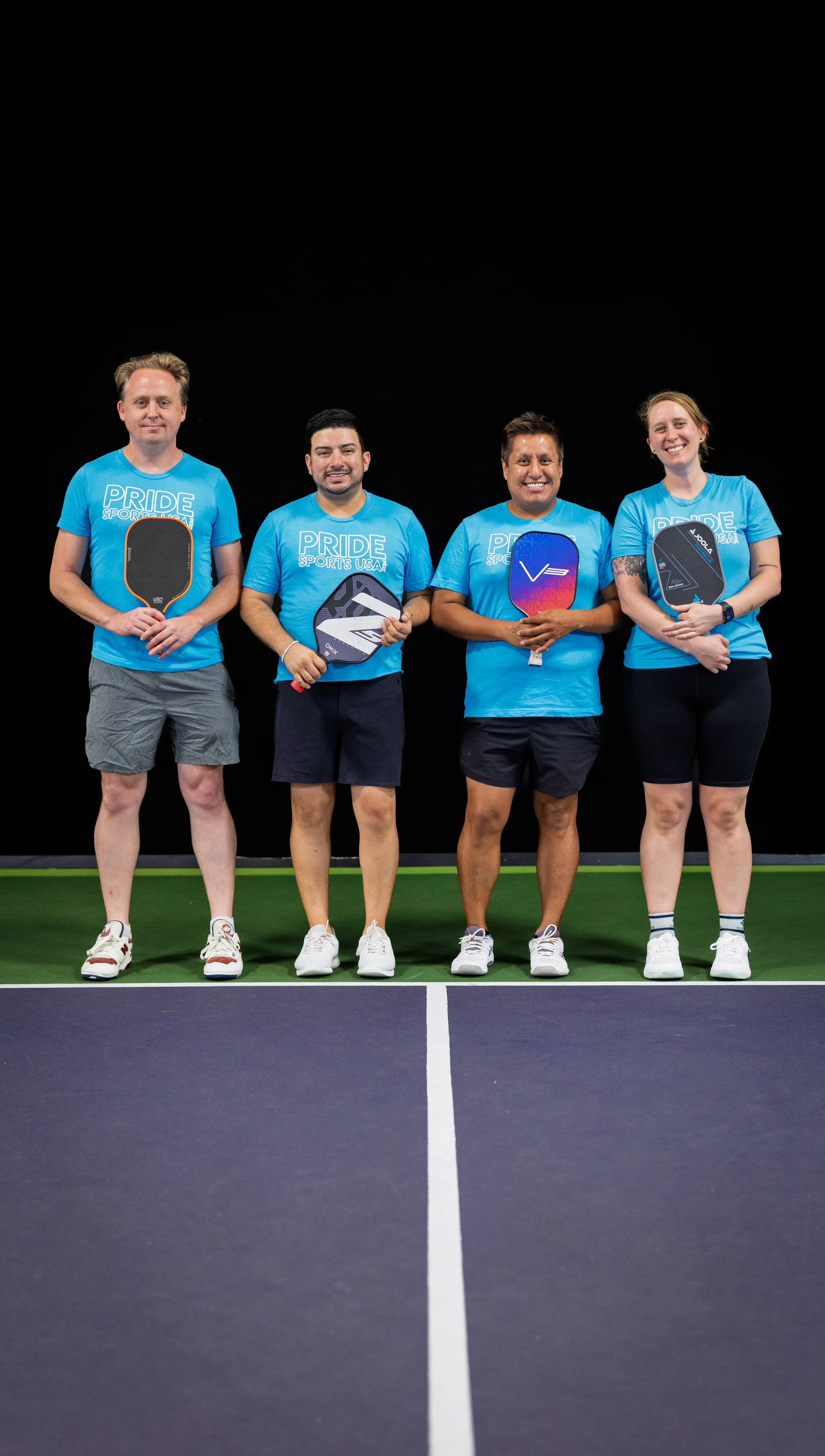 Four pickleball players stand side by side on a court holding paddles—black, gray-and-white zigzag, blue-purple, and black with red accents—wearing matching bright blue “PRIDE” shirts, dark shorts, and athletic shoes. They pose near the centerline against a dark backdrop, looking ready for a Summer 2025 match.