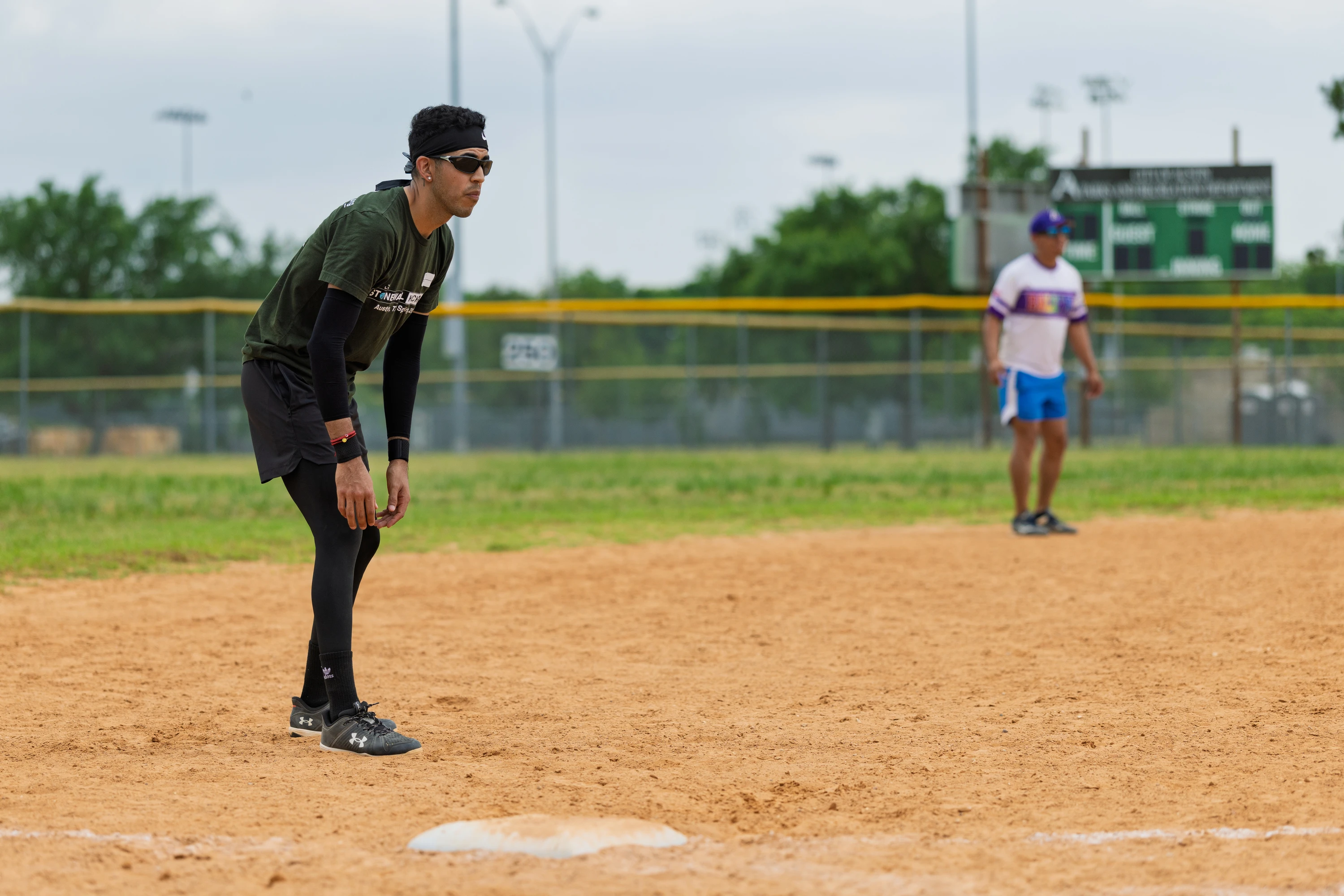 A player in a black headband, dark shirt, and leggings is positioned alertly near a base on a kickball field, while another person stands in the outfield wearing a white and purple shirt and blue shorts.