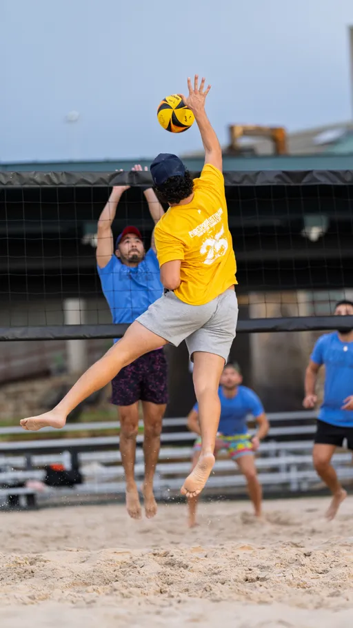 A volleyball player in a yellow shirt and cap leaps to spike the ball over the net, while an opposing player in a blue shirt and hat jumps to block; other teammates stand ready on the sandy outdoor court.