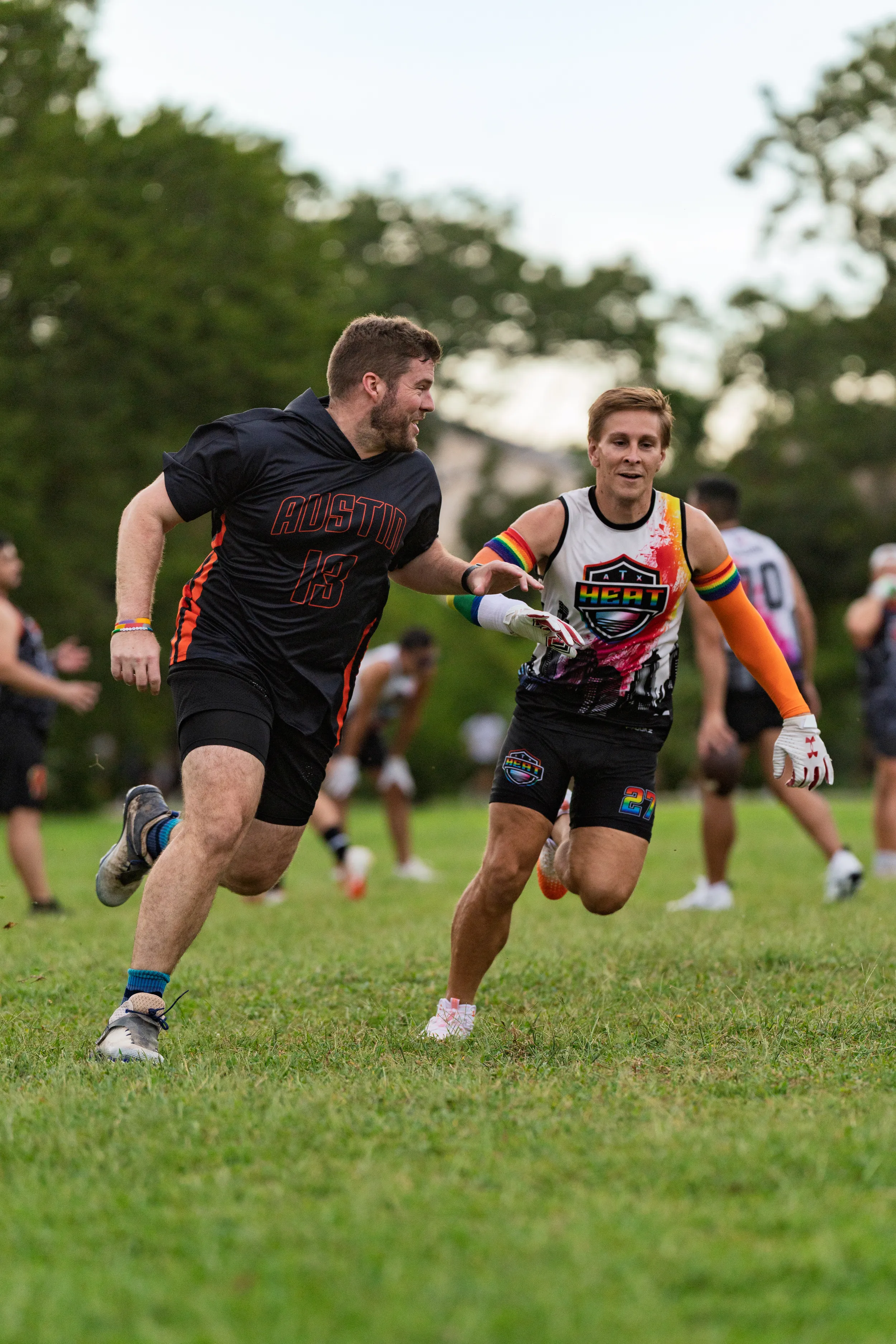 Two ATX Heat (Orange) players sprint during flag football practice for Gay Bowl 2025, with one in a black “AUSTIN 13” kit running upfield while a defender in a white-and-orange sleeveless jersey, gloves, and an arm sleeve reaches to make a tag. Teammates move in the background on a grassy field bordered by trees.
