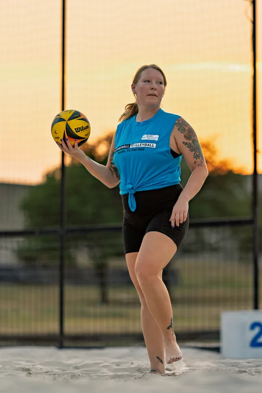 A player on a sand volleyball court at sunset holds a yellow-and-black Wilson ball, poised to serve. Wearing a sleeveless blue Stonewall Volleyball shirt tied at the waist and black shorts, the athlete stands barefoot inside a fenced court with a visible court marker.