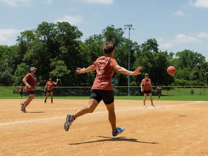 A player wearing a red team shirt and black shorts is in mid-stride after kicking a red kickball on a dirt kickball field, with several other players in similar uniforms positioned around the field under a sunny sky.
