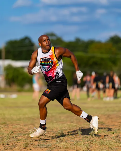 An ATX Heat (Black) player runs on a grassy flag football field during Gay Bowl practice, wearing a multicolor Heat jersey, black shorts with “21,” white gloves, and white cleats. The athlete looks upfield with arms ready as teammates move in the blurred background under a bright sky.