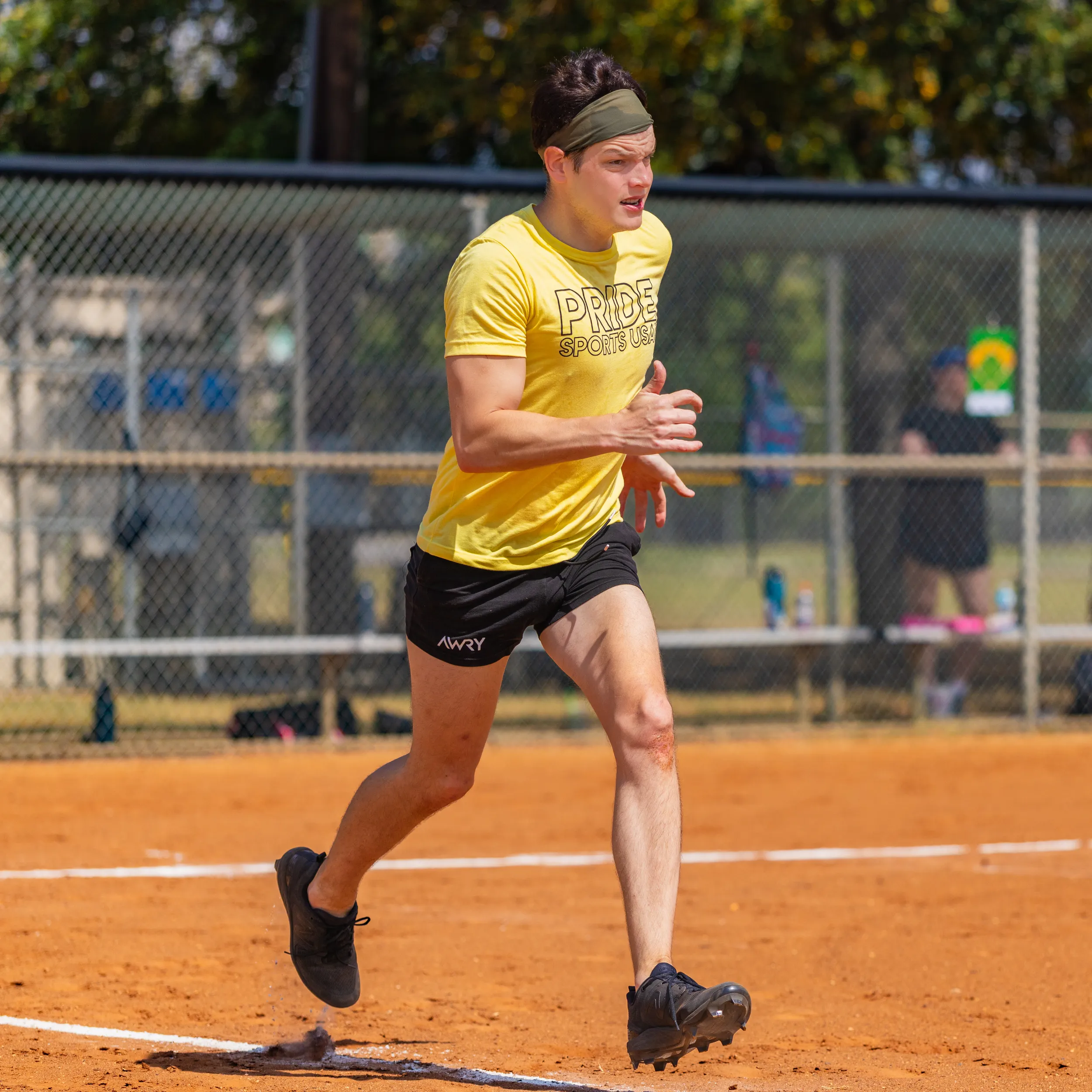 A player wearing a yellow “PRIDE SPORTS” shirt, black shorts, cleats, and a headband sprints on the red-dirt kickball field, kicking up dust as they cross a base line. The scene is a sunny park with a chain-link fence and blurred teammates and gear in the background.