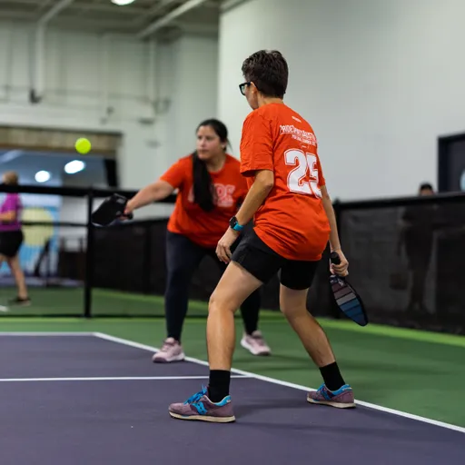 Two pickleball players wearing matching red shirts and athletic gear are engaged in action on an indoor court, with one player moving forward to hit an incoming ball with a paddle while the other stands ready nearby.