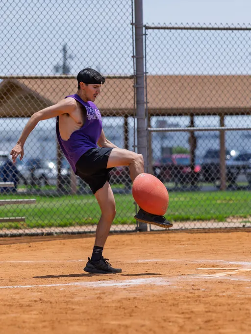 A player wearing a purple sleeveless shirt and black shorts is kicking a red kickball mid-air on a dirt kickball field, with a chain-link fence and grassy area in the background.